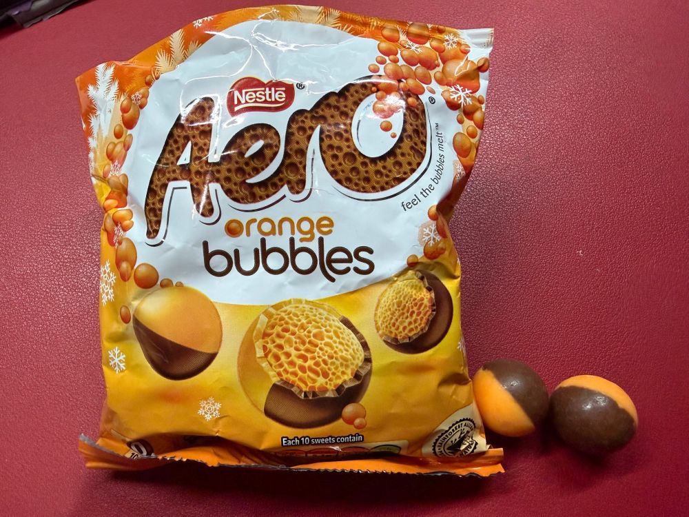 photo of a a packet of Aero Orange bubbles on a red background.  there is a picture showing one of the chocolates cut in half which shows orange bubbles.  next to the bag on the right are two chocolate balls.  each ball is half orange and half milk chocolate