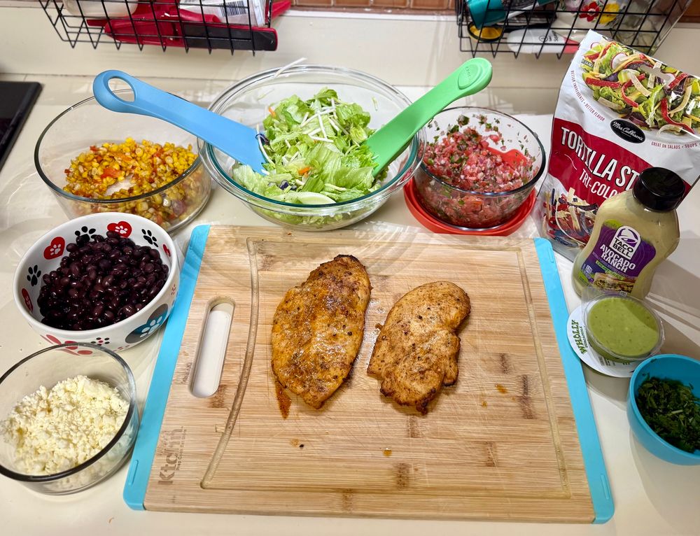 2 thin honey and lime chicken breasts on a cutting board with bowls of cojita cheese, black beans, corn, lettuce, and salsa around the left side and back, with a bag of tortilla strips, avocado ranch dressing, guacamole and cilantro on the right side. 