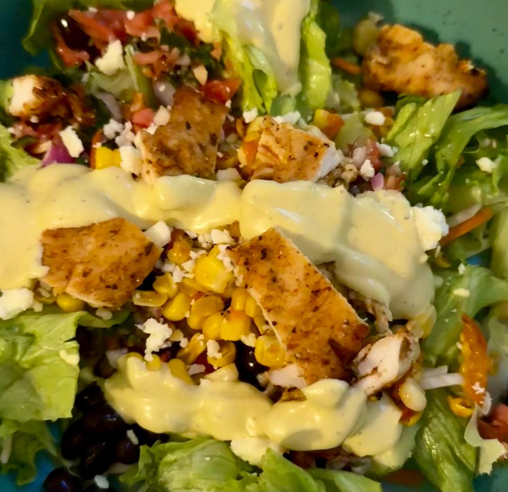 A salad in a bowel with lettuce, black beans, corn, salsa, cojita cheese, chicken breast, and avocado ranch dressing. 
