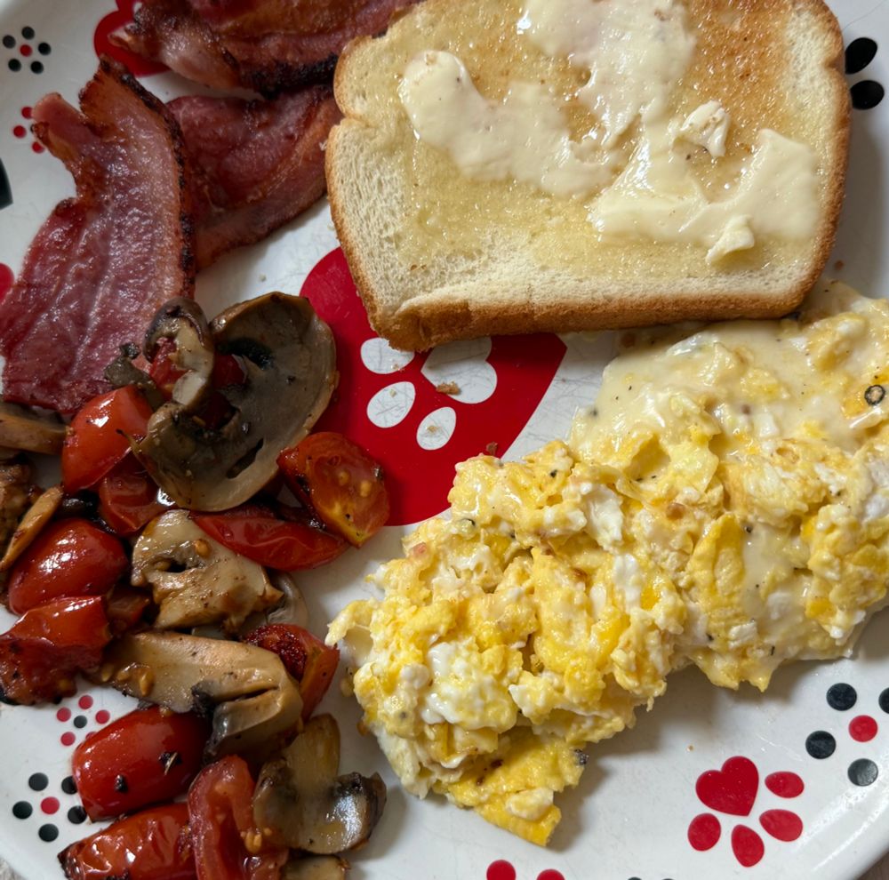 3 half pieces of bacon, above a pile of cooked tomatoes and mushrooms, next to scrambled eggs, with a piece of buttered toast above, on a plate. 