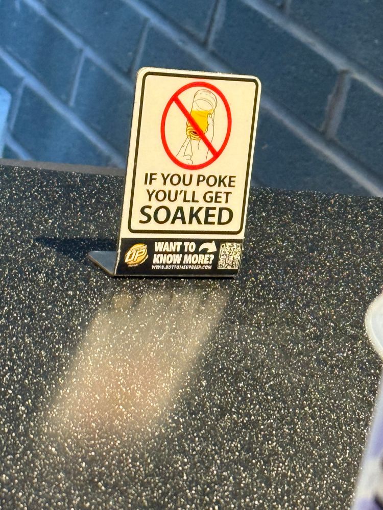 A sign at the bar that says “IF YOU POKE YOU'LL GET SOAKED
WANT TO A KNOW MORE?
WWW.BOYTOMSOPBARA.COM” with illustration of someone poking the bottom of a cup with a red circle and slash over it. 