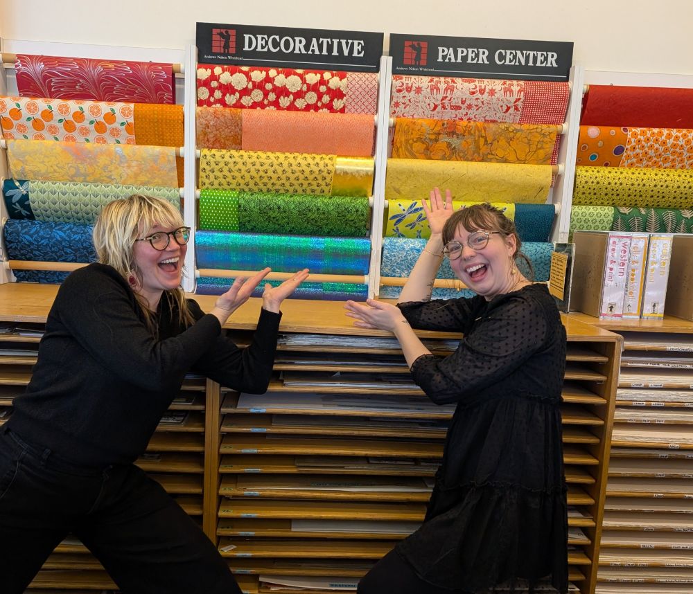 Two Wet Paint employees standing in front of the paper display in the Paper Department at the store.