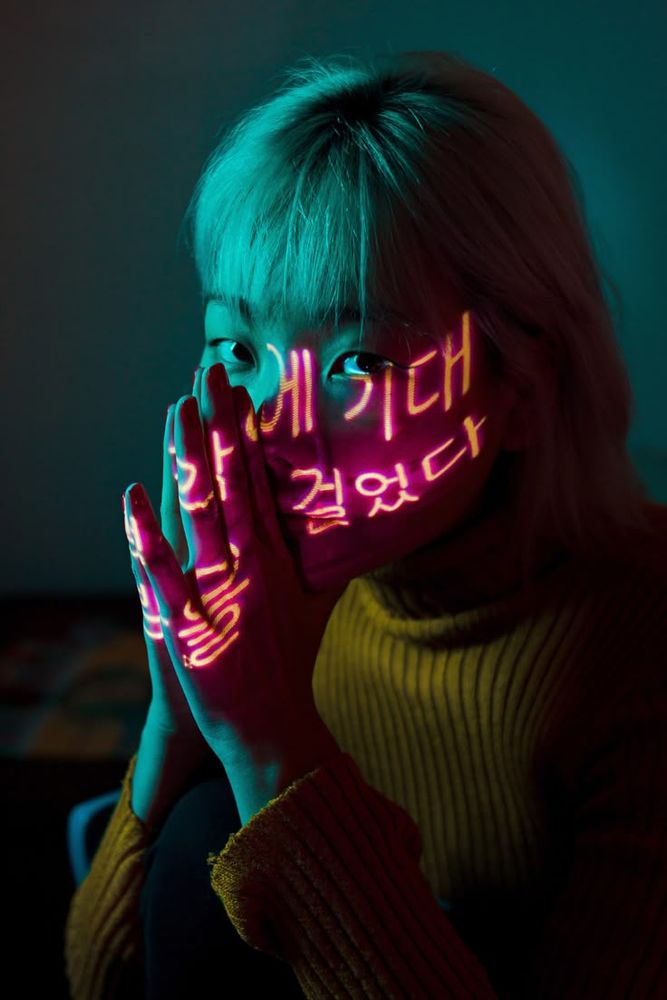 Lighting reference of girl, holding hands close to her face, in teal and redish pink lighting with korean lettering lighting left side of face.