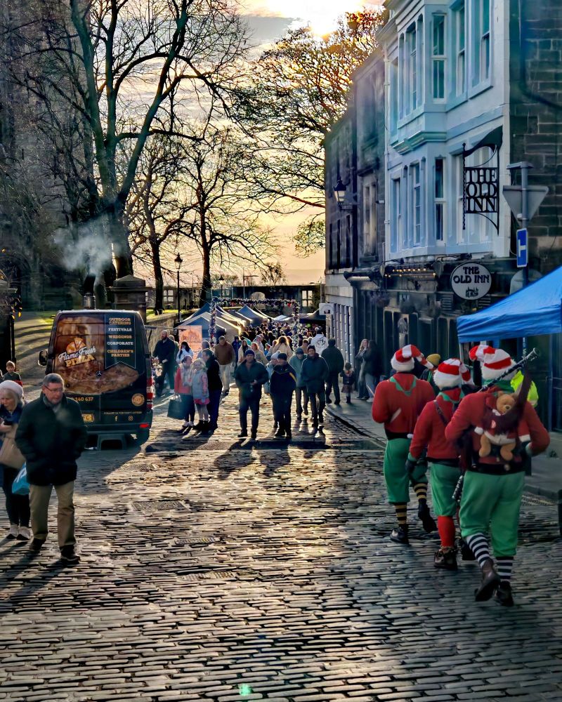 In the winter sunshine 3 men dressed as elves head down the cobbled stones past The Old Inn pub towards Dunfermline Abbey with a market taking place. 