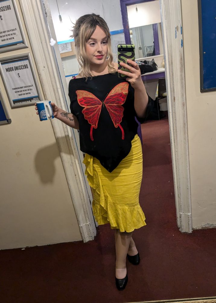 A full length mirror selfie of me (Toni) wearing a blonde wig, a black top with a large butterfly applique, and a bright yellow 3/4 length skirt. The outfit is a recreation of Anya Jenkins outfit during 'I Gotta Theory/Bunnies/What Can't We Face' from the musical episode of Buffy the Vampire Slayer, Once More With Feeling.
