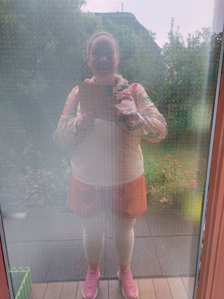 Spiegelselfie in Glastür. Ich trage pinke Schuhe, gelbe Socken, orange kurze Hose unter beiger Jacke mit bunten Mustern. Hinter mir ein Garten mit Rosen