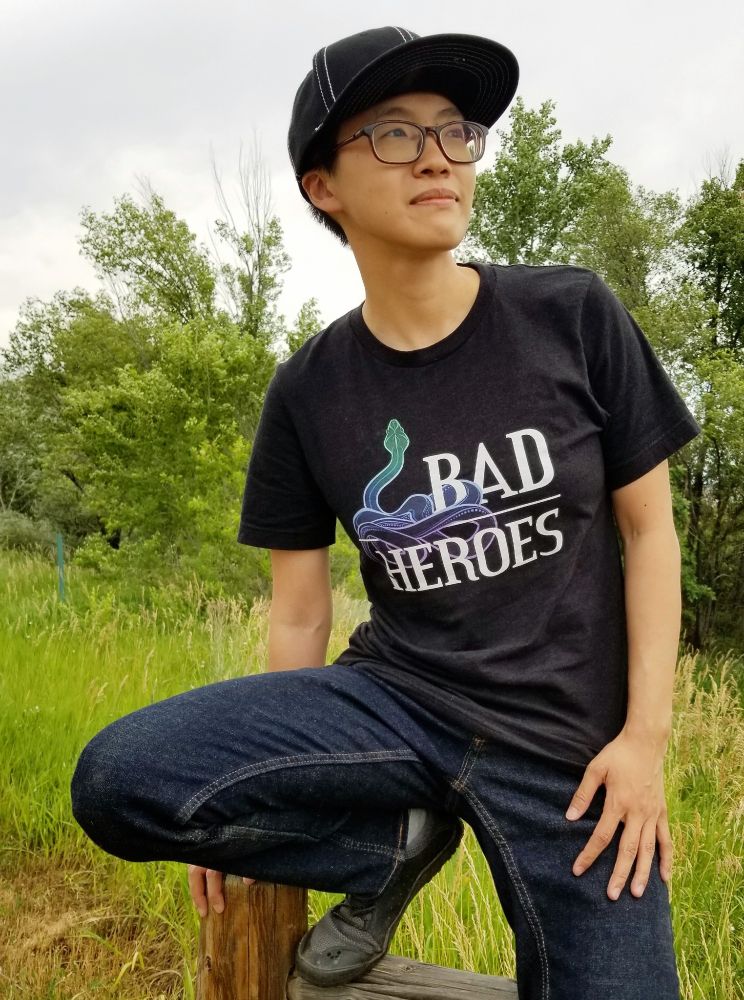 Photo of Lian Xia Rose, an East Asian person perched on a low wooden fence and looking to the right, wearing a black snapback hat, glasses, jeans, sneakers and a black t-shirt with the Bad Heroes snake logo on the front. There is a grassy field and trees in the background.