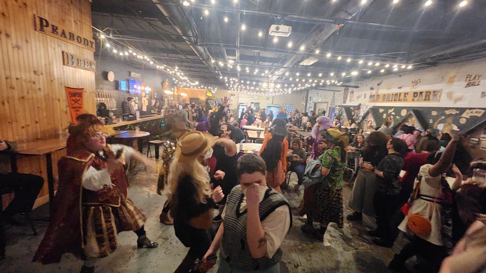 People in costume dance in a crowded brewery. A jester, a person in a long Wig and hat, Gritty, a drag queen in purple sequins, and a person dressed as a Persian royal are visible. The jester is throwing it down and looks cool as fuck.