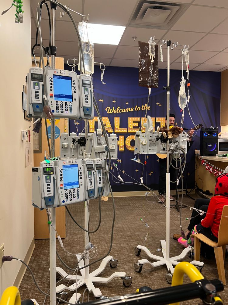A hospital family lounge space with a full wall sticker reading “Welcome to the TALENT SHOW” in yellow on a dark blue background. A man is playing guitar in front of the wall, but can hardly be seen behind 3 IV poles.