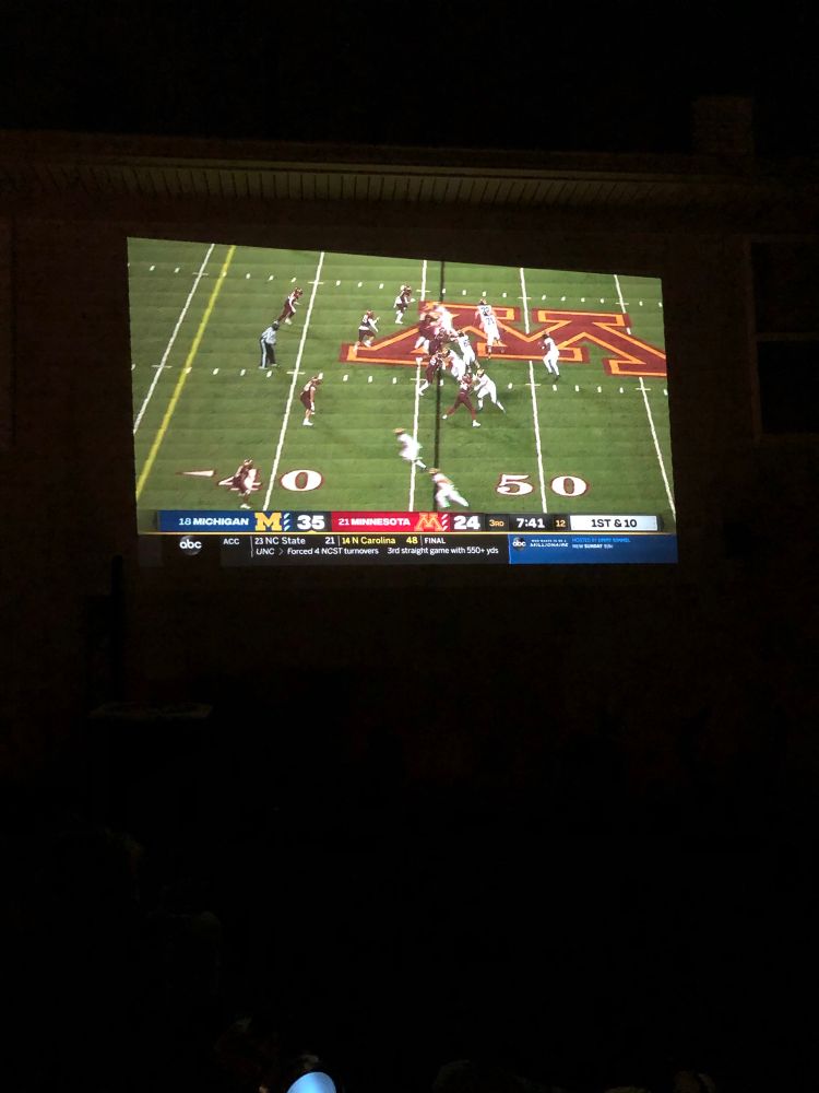 Michigan-Minnesota football (2020 edition) projected onto the side of a house. It’s dark out so it’s hard to see the house, it mostly just looks like the football.