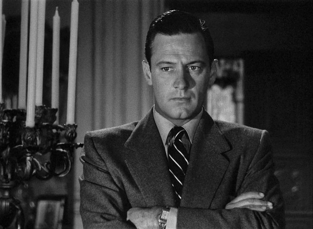 William Holden as Joe Gillis standing in a suit with his arms crossed over his chest.