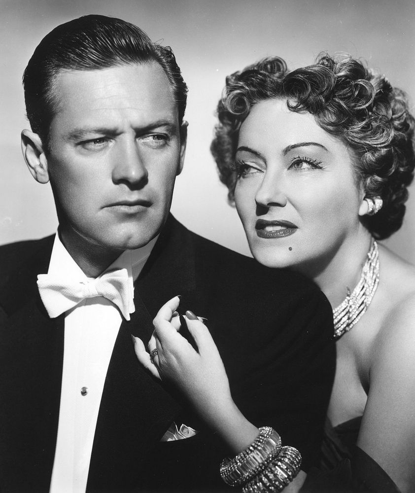 A medium close up of William Holden as Joe Gillis frowning off to the side. Norma Desmond (Gloria Swanson) is just behind him and her hand is reaching around to his chest.