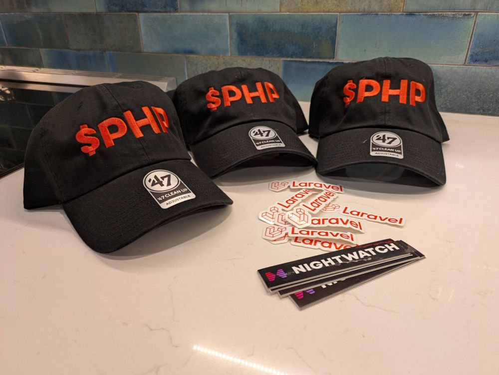 Three black hats sit on a counter next to a pile of stickers. The hats say PHP in red letters, the stickers say Laravel and Night watch.