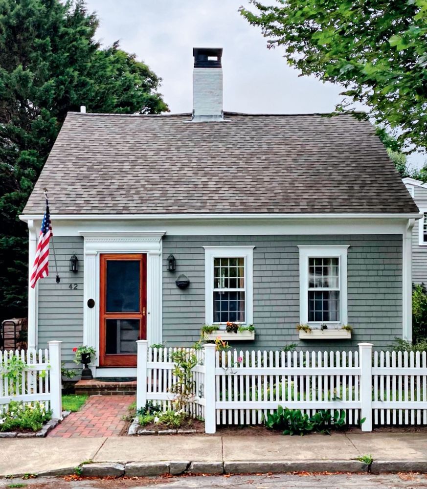 A classic old Cape Cod cottage with muted grey siding, white trim, and a red door.
