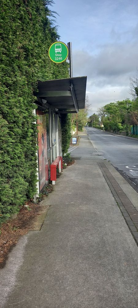 A bus shelter on Rochestown Avenue in Dublin. The footpath is narrow so the back of the bus shelter is right up against the stone boundary wall of a building and the roof juts out from there,  The seating is also very narrow. 