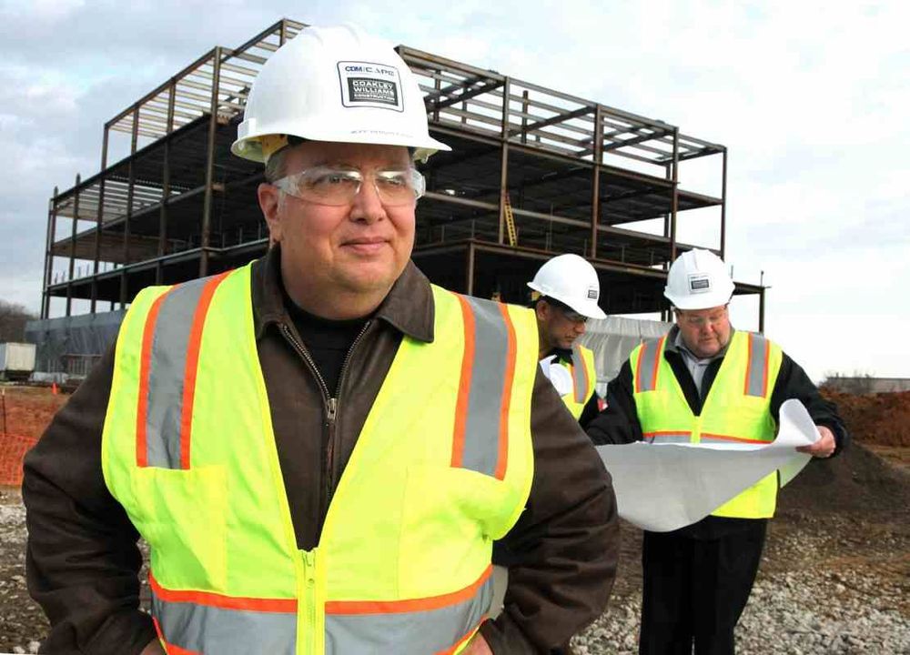 Columbia MD portrait photography of architect on building construction site in PPE safety clothing and hardhat; a group of people at work collaborating over industrial drawings is behind him with a partly-built steel warehouse/