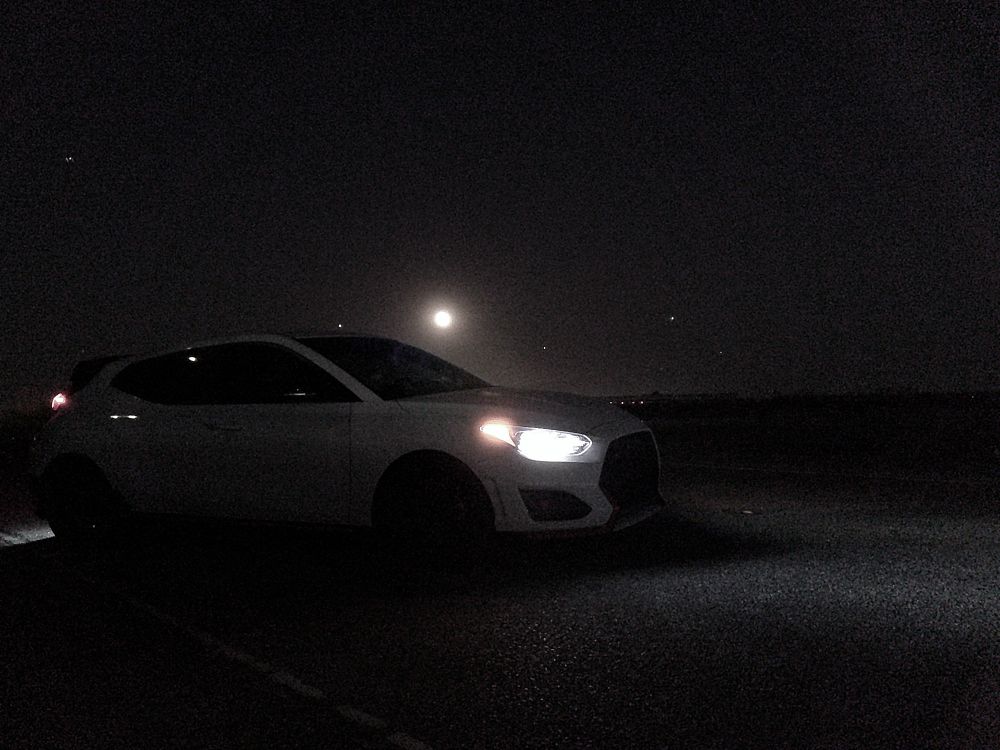 The large moon can be seen as a white dot across a black sky above a Veloster N parked in the road.