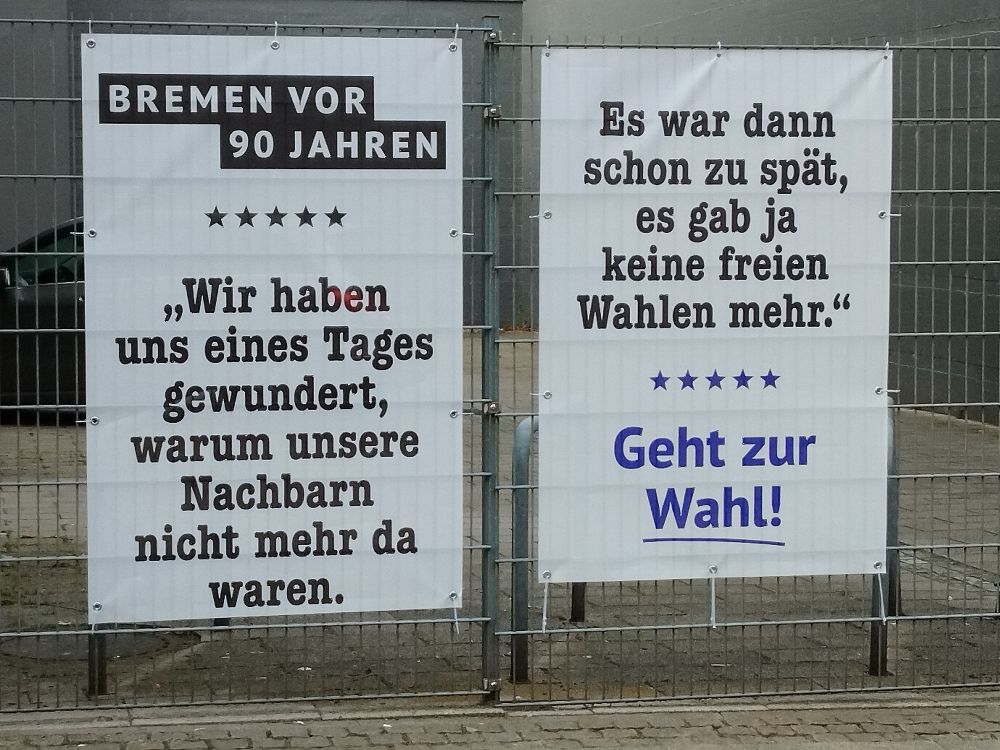 Das Bild zeigt zwei Transparente an einem Zaun. Auf ihnen steht: Bremen vor 90 Jahren: "Wir haben uns eines Tages gewundert, warum unsere Nachbarn nicht mehr da waren. Es war dann schon zu spät. Es gab ja keine freien Wahlen mehr." Geht zur Wahl!