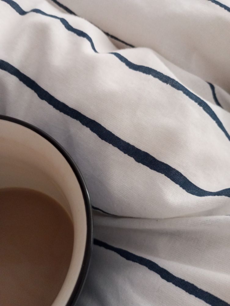 Bild zeigt fast leeren Tasse mit Milchkaffee auf einer weißen Bettdecke mit blauen Streifen.