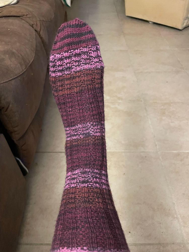 A hand knit sock in stripes of shades of purple, pink, and orange-red with a black undertone. 
