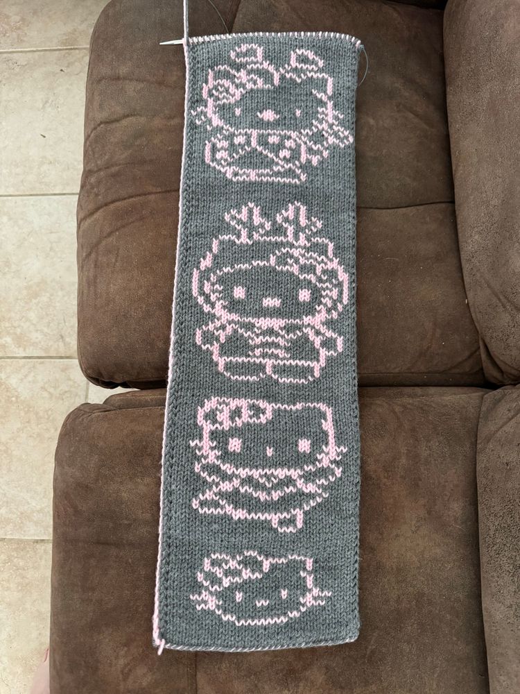 The reverse side of a double knit scarf with a dark gray background and dark pink outlines showing, from top to bottom, Hello Kitty as a ladybug, Hello Kitty wearing a hood with antlers, Hello Kitty dancing, and a basic Hello Kitty face. 