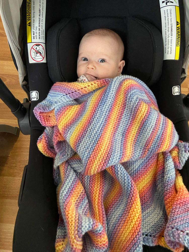 A very cute baby snuggled up under a hand-knit garter stitch baby blanket made of self-striping yarn in pastel multicolored shades. 