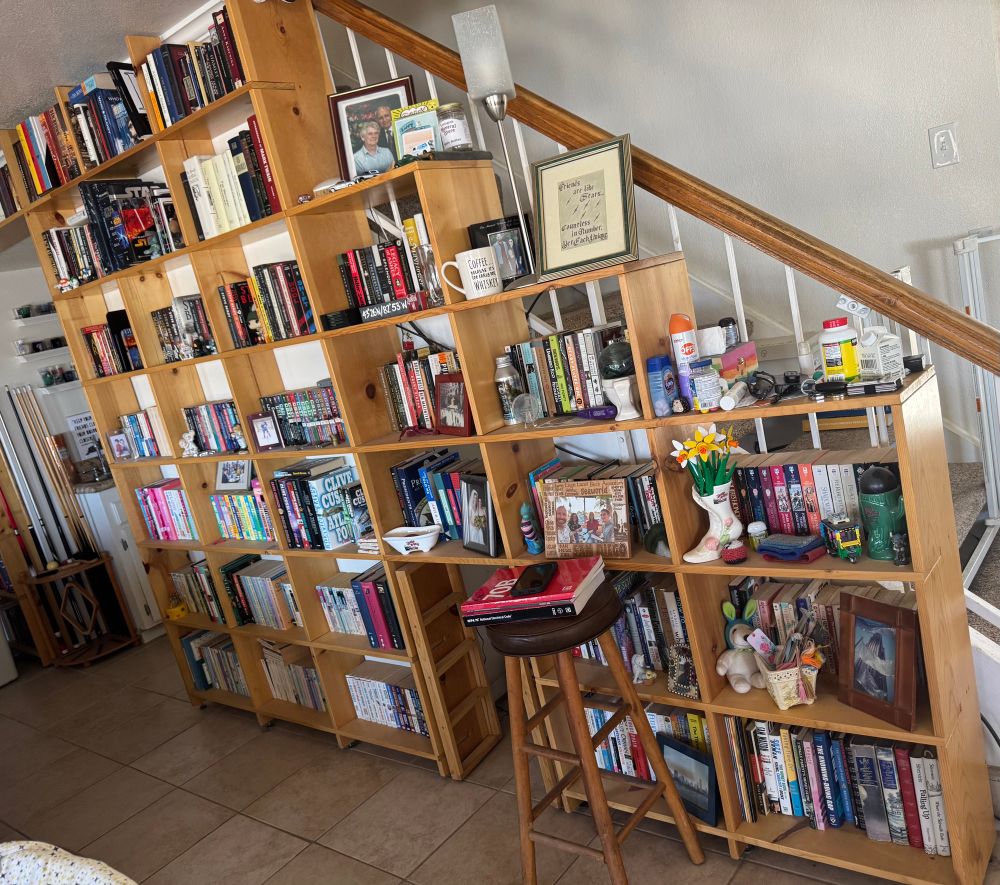 An angled view of custom built bookcases that follow the profile of a staircase, filled with books, photos, and knickknacks. In front of the bookcase is a stool with electrical safety code books sitting on top. 