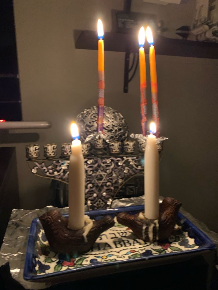 Hanukkiah with the Shamash and two candles lit. Candles are orange on the top 1/3 and the bottom 2/3 is multicolored. Two white Shabbat candles in holders that look like birds are on a tray in the foreground