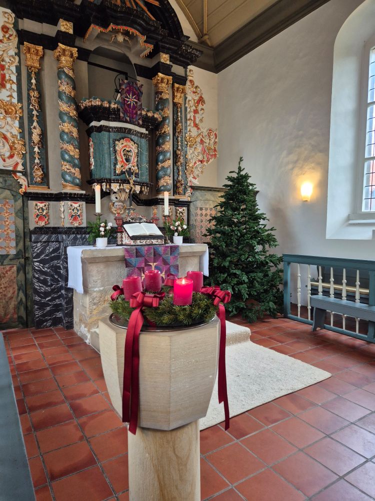 Eine rote Kerze brennt an einem Adventskranz mit roten Kerzen und roten Schleifen im Altarraum einer Kirche. Im Hintergrund steht eine ungeschmückte Tanne.