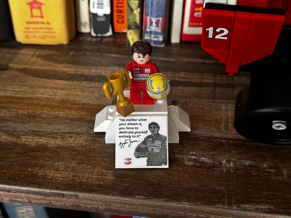 A Lego minifigure of Ayrton Senna holding a trophy and his signature yellow helmet, standing on a pedestal. The pedestal reads “No matter what your dream is, you have to dedicate yourself entirely to it.”
