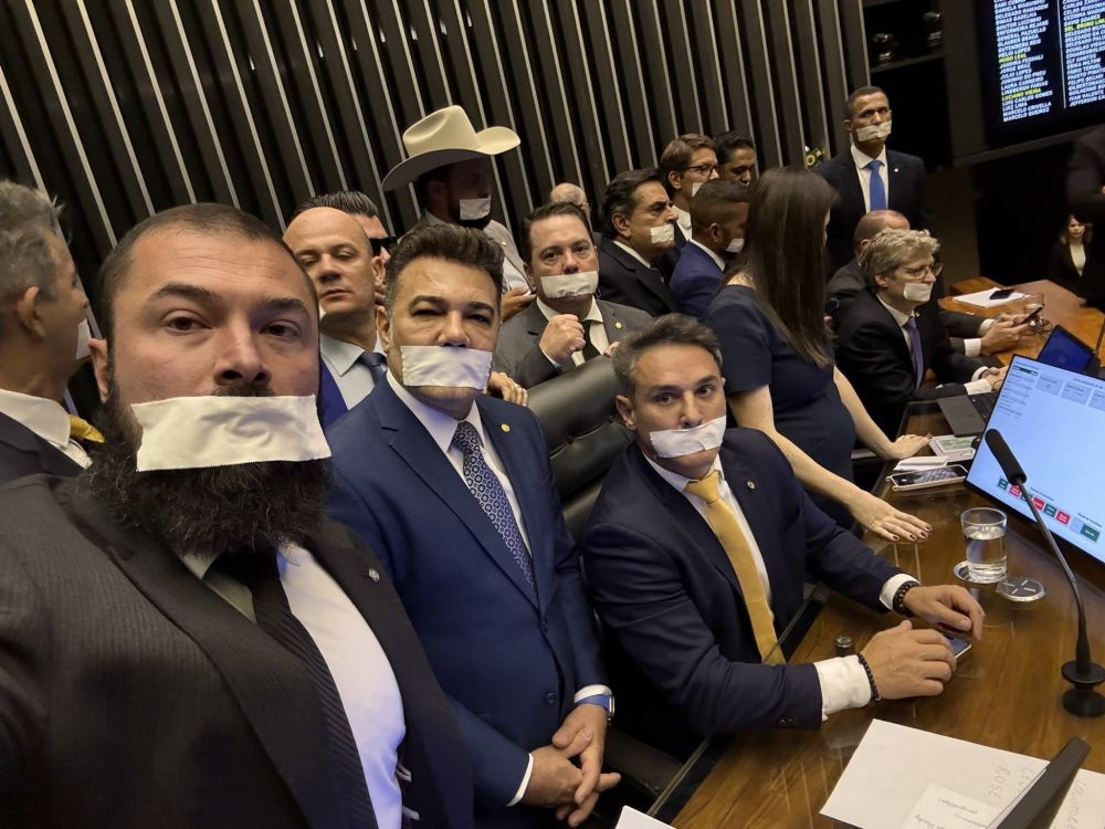 A imagem mostra um grupo de homens, a maioria vestindo terno e gravata, dentro de um ambiente que aparenta ser um plenário ou uma sala de comissão de um órgão legislativo. Vários deles, incluindo o que está em primeiro plano tirando a foto (selfie), estão com as bocas cobertas por um esparadrapo ou fita branca. Eles estão sentados em suas bancadas, e a expressão coletiva sugere um ato de protesto silencioso, simbolizando censura ou a impossibilidade de falar. Ao fundo, um homem usa um chapéu de cowboy.

A imagem é uma selfie tirada pelo deputado federal Gustavo Gayer (PL-GO), que aparece em primeiro plano com a boca coberta por uma fita branca. A foto foi registrada no plenário da Câmara dos Deputados e mostra outros parlamentares da oposição também amordaçados em sinal de protesto.

Atrás de Gayer, são visíveis outros deputados, incluindo o Coronel Zucco (PL-RS), de terno azul no centro da imagem, e Rodolfo Nogueira (PL-MS), que está sentado à sua mesa usando terno escuro e gravata amarela. Ao fundo, um dos participantes do ato usa um chapéu de cowboy.