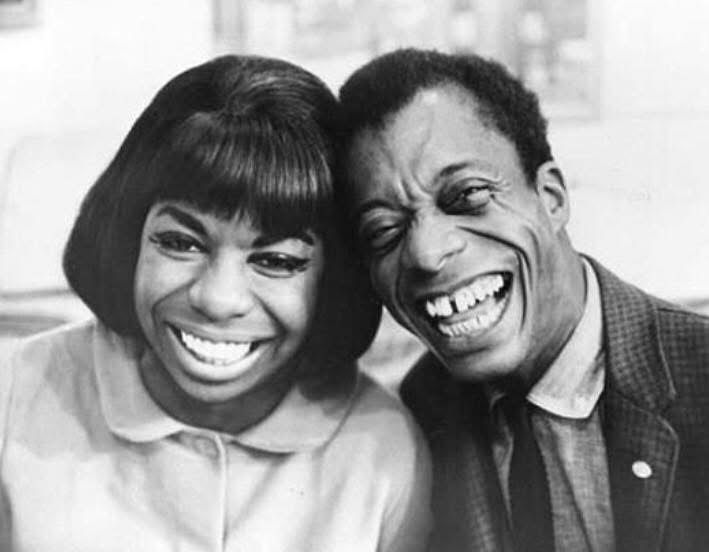 The image captures a moment between American author and activist James Baldwin and American singer, songwriter, and civil rights activist Nina Simone. 

The photograph, taken by Bernard Gotfryd in 1964, shows Baldwin and Simone smiling joyfully toward the camera. Ms. Simone, on the left, is dressed in a white, button-down round-collared, blouse; Baldwin in a dark sport jacket, white shirt and thin, black necktie.
