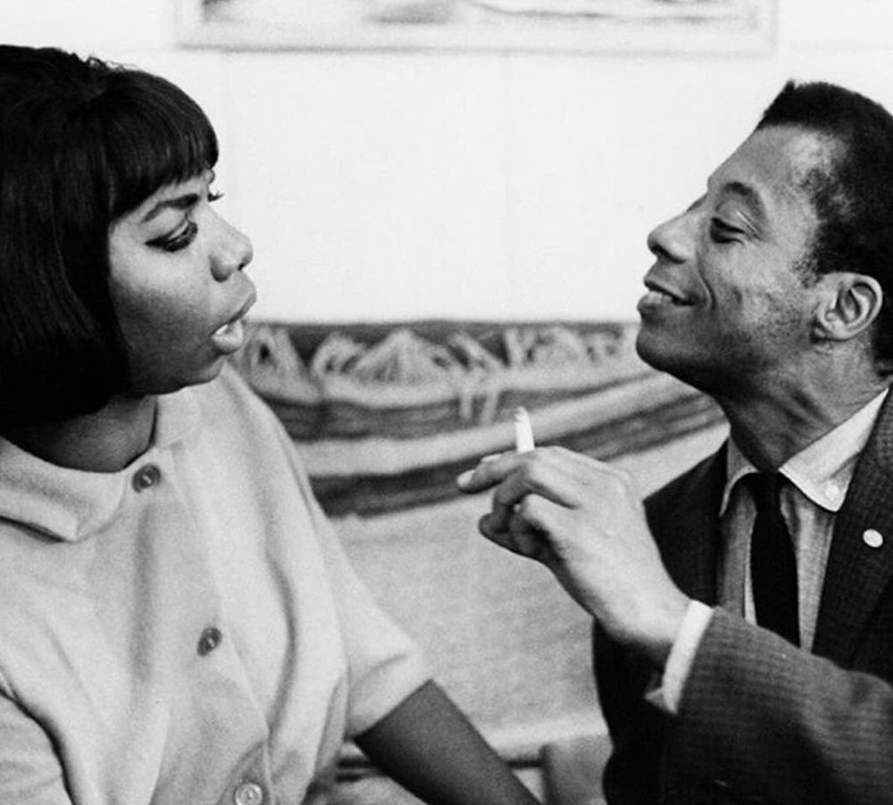 The image captures a moment between American author and activist James Baldwin and American singer, songwriter, and civil rights activist Nina Simone. 

The photograph, taken by Bernard Gotfryd in 1964, shows Baldwin and Simone engaged in friendly conversation. Ms. Simone, on the left, is dressed in a white, button-down round-collared, blouse; Baldwin in a dark sport jacket, white shirt and thin, black necktie.