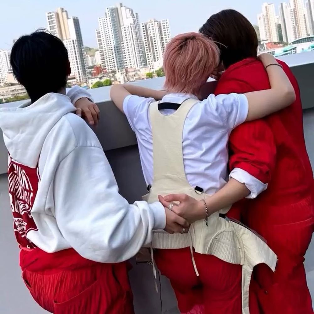 Myung Jaehyun, Riwoo, and Leehan of BoyNextDoor. Riwoo has his arm on Leehan's shoulder and Jaehyun and Leehan are holding hands behind him.