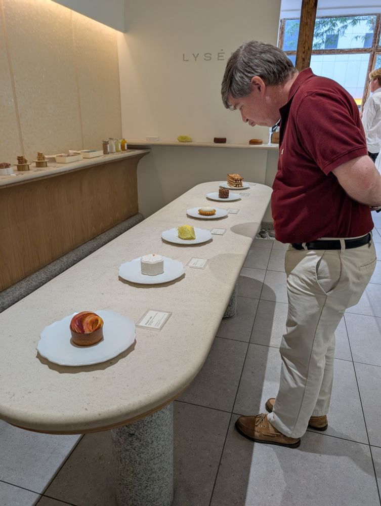 M in the gallery of Lysée, looking closely at the desserts on offer to purchase. The desserts are arranged on a long table in the center and a shelf along the wall. We are asked to not touch the desserts.