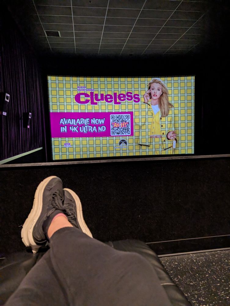Clueless in a movie theatre. My feet are up in the reclining chair. I'm wearing black sweats and sneakers. You can't see the rest of the theatre but it is filled with women my age all stoked to be here