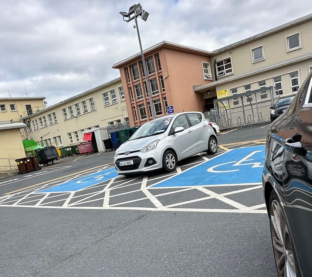 A silver Hyundai i10 (I think) parked in the space BETWEEN two blue badge spots (an exclusion zone marked with hatching to allow space for people with restricted movement or who might need equipment to mobilise). The car is plastered in both Learner and Novice plates, but not a blue badge to be seen. The car park in question is part of the Louth Hospital complex in Dundalk, Co. Louth, which makes the infraction even worse, if anything (plus there were plenty of free spaces available).
