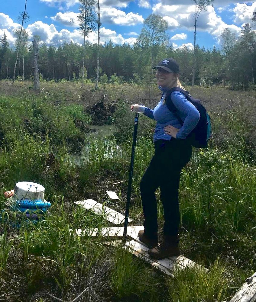 Laura, wearing a black ball cap, light blue long-sleeved top, black trousers, and hiking boots, is standing on a boardwalk in a peatland and smiling at the camera. There are trees in the background. Behind Laura is a pool, and to the left are a couple of floating chambers stacked up which are used to measure greenhouse gas fluxes from peatland waterbodies.