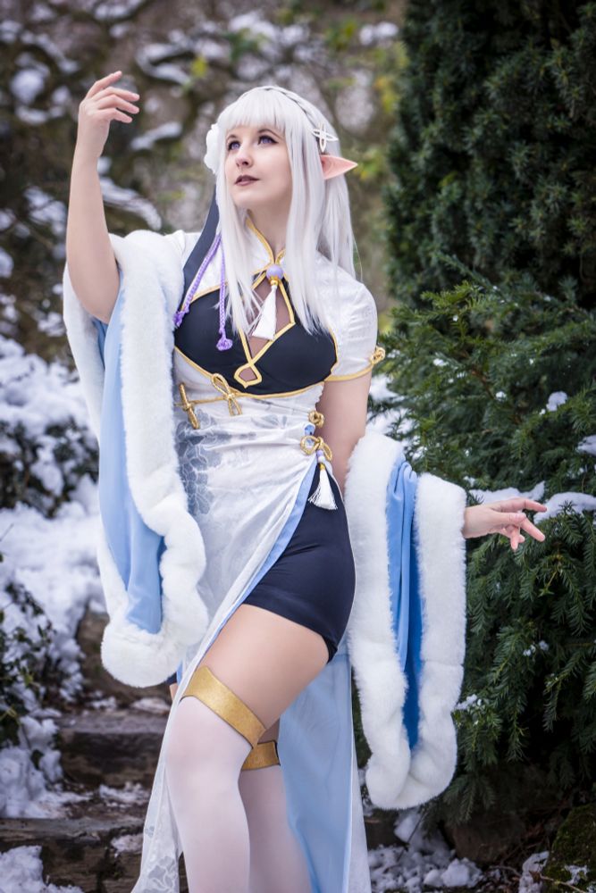 Eine junge Frau mit langen, weißen Haaren und spitzen Elfenohren steht anmutig in einer verschneiten Landschaft. Sie trägt ein aufwendig gestaltetes Cosplay-Kostüm mit asiatischen und Fantasy-Elementen: ein weiß-blaues Kleid mit goldenen Details, einen pelzbesetzten Mantel und weiße Strümpfe mit goldenen Bändern. Ihr Blick ist verträumt nach oben gerichtet, während sie eine Hand in die Luft hebt, als würde sie Magie wirken. Der Hintergrund zeigt eine winterliche Naturkulisse mit schneebedeckten Steinen und grünen Nadelbäumen.
