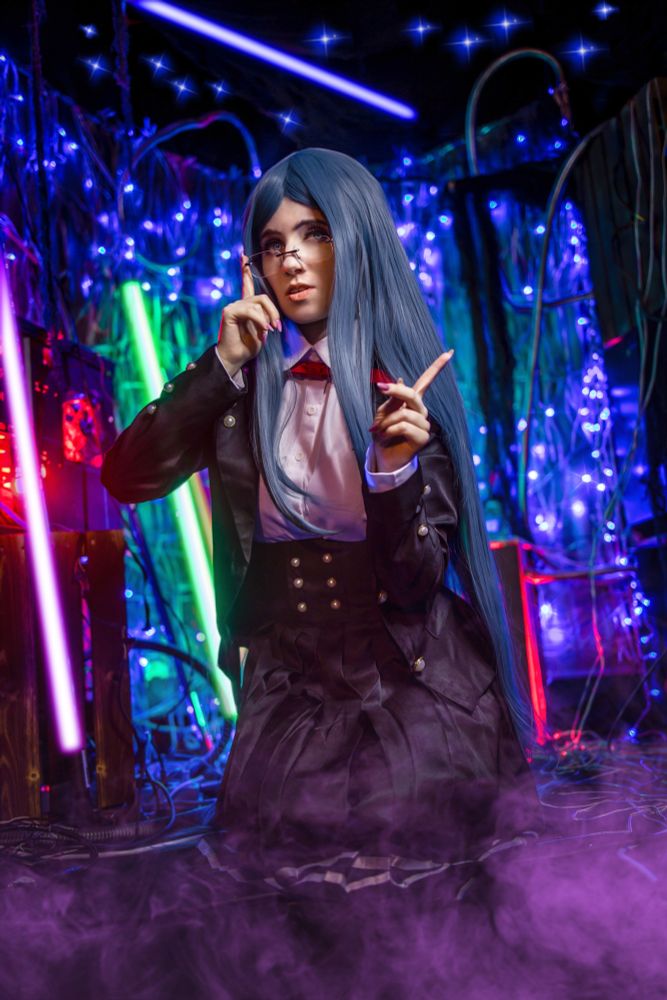 Eine Cosplayerin stellt Tsumugi Shirogane aus dem Videospiel „Danganronpa V3: Killing Harmony“ dar. Sie trägt eine lange blaue Perücke, eine runde Brille, ein weißes Hemd mit roter Schleife und eine schwarze Uniform mit silbernen Knöpfen. Sie hebt mit einer Hand ihre Brille an und zeigt mit der anderen Hand nach oben. Der Hintergrund ist bunt und futuristisch mit leuchtenden Neonröhren in Grün, Pink und Blau sowie vielen glitzernden Lichtern und sichtbaren Kabeln. Am Boden wabert violetter Nebel.