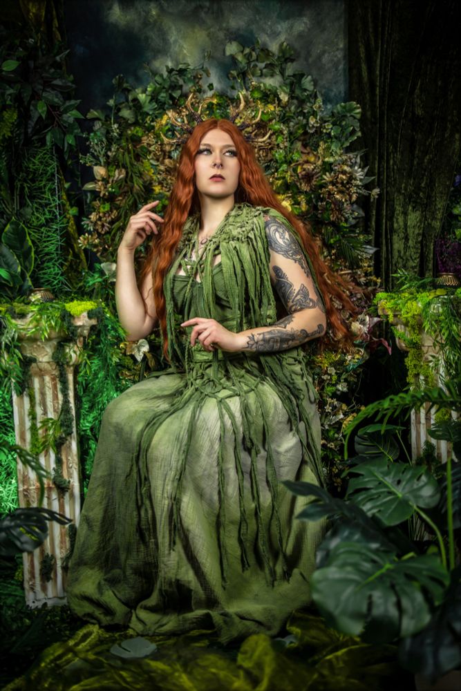 Eine Cosplayerin oder Fantasy-Darstellerin sitzt auf einem kunstvoll dekorierten Thron aus Pflanzen und Blumen. Sie trägt ein langes, moosgrünes Kleid mit fransigen Stoffstreifen und hat langes, rotbraunes Haar. Ihr Kopf wird von einem filigranen Geweih-Kopfschmuck gekrönt. Ihr linker Arm zeigt aufwendige schwarze Tätowierungen. Die Szene erinnert an eine Naturgöttin oder Dryade, umgeben von üppigem Grün, Blättern und antiken Säulen, überwuchert von Moos.
