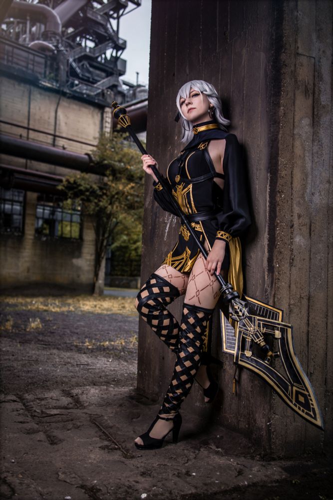Eine Cosplayerin stellt Noelle aus dem Mobile-Game NieR Re[in]carnation dar, in ihrem eleganten schwarzen und goldenen Celebratory-Weapon-Outfit. Sie lehnt lässig an einer Betonwand in einem Industriegebiet mit alten Rohren und Gebäuden im Hintergrund. Das Kostüm besteht aus einem schwarzen Einteiler mit goldenen Ornamenten, hohen geschnürten Beinschienen und einem langen gelben Stoffelement am Rücken. In der Hand hält sie eine große, kunstvoll verzierte Hellebarde mit geometrischem Design. Ihr silbernes Haar fällt locker über die Schultern, der Blick ist selbstbewusst nach vorn gerichtet.