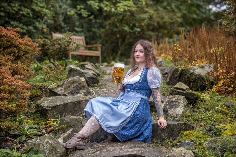 Eine Frau mit welligem, schulterlangem Haar sitzt entspannt auf einer Felslandschaft inmitten eines üppigen, grünen Gartens. Sie trägt ein traditionelles bayerisches Dirndl in Blau mit weißen Spitzenärmeln und hält eine Maß Bier in der Hand. Ihre Beine sind tätowiert, und sie trägt braune Lederstiefel. Im Hintergrund steht eine Holzbank, umgeben von herbstlich gefärbten Pflanzen. Sie blickt mit einem zufriedenen Lächeln in die Kamera, während das goldene Bier im Glas schimmert.