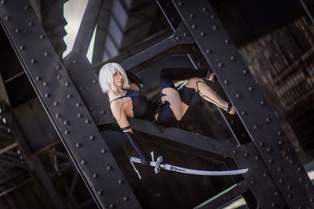 Eine Cosplayerin mit platinblonder Kurzhaarperücke posiert lässig in einem schwarzen, futuristischen Outfit in einem stählernen Industriegelände. Sie liegt in einem diagonalen Stahlträgergerüst, den Blick selbstbewusst zur Seite gerichtet. In der Hand hält sie ein langes Katana mit silberner Klinge. Das Setting wirkt urban und kontrastiert mit dem eleganten, kampfbereiten Look der Figur.