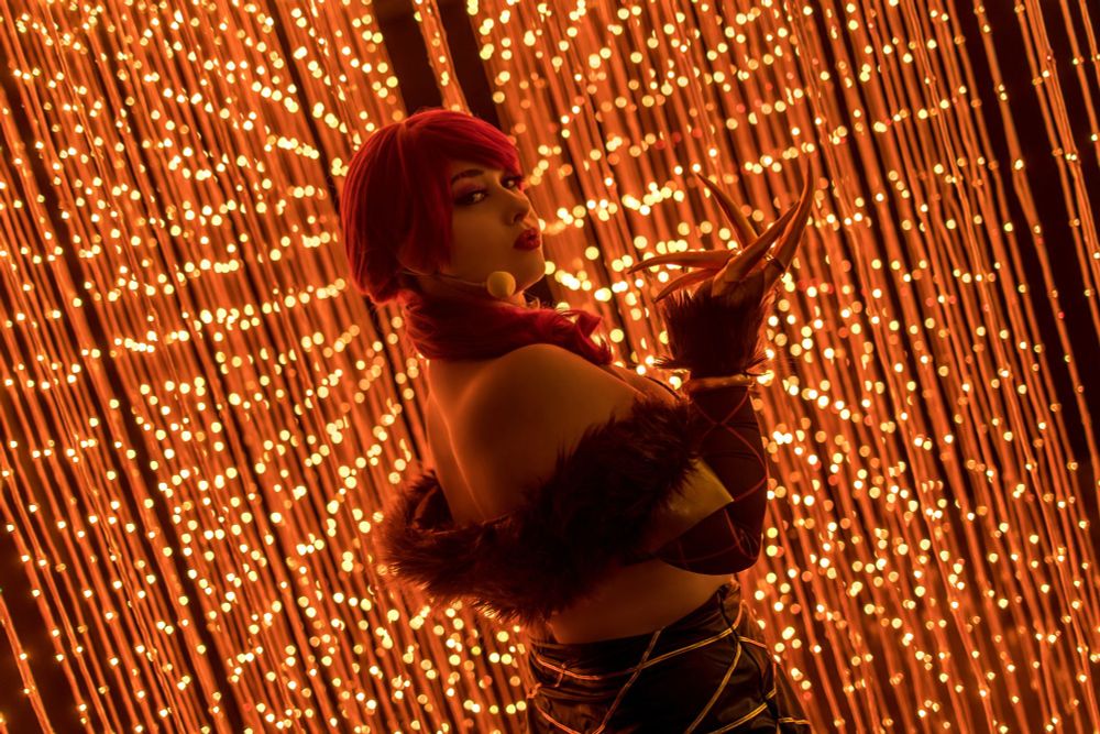 Eine Person in einem auffälligen Fantasy-Kostüm posiert vor einem funkelnden Hintergrund aus goldenen Lichterketten. Sie hat leuchtend rote Haare, die in einen eleganten Zopf gelegt sind, und trägt ein Outfit mit Fellbesatz, langen Handschuhen und auffälligen goldenen Krallen an den Fingern. Ihr Blick ist intensiv, mit dunklem Make-up und tiefroten Lippen. Die Szene strahlt eine geheimnisvolle, glamouröse Atmosphäre aus, während die warmen, goldenen Lichter hinter ihr eine magische Kulisse bilden.