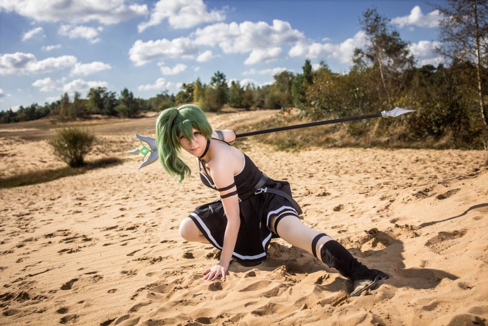 Eine Person im Cosplay posiert kämpferisch in einer weitläufigen Sandlandschaft bei sonnigem Wetter. Sie trägt ein schwarzes, schulterfreies Outfit mit weißen Zierlinien und schwarze Stiefel. Auffällig ist die grüne Perücke mit seitlichem Pferdeschwanz. Die Cosplayerin hält einen langen, dekorativen Speer, der über ihre Schultern gelegt ist. Im Hintergrund sind Bäume und blauer Himmel mit weißen Wolken zu sehen.