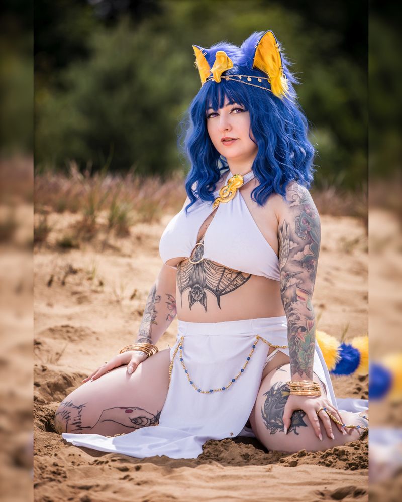 Eine Person im fantasievollen Cosplay sitzt im Sand einer natürlichen Dünenlandschaft. Sie trägt ein weißes, bauchfreies Outfit mit goldenen und blauen Schmuckelementen. Auffällig sind die blaue Perücke mit großen, flauschigen Katzenohren in Blau und Gelb sowie die vielen sichtbaren Tattoos an Armen, Bauch und Beinen, darunter ein großes Fledermaus-Tattoo unter dem Brustbereich. Die Cosplayerin blickt selbstbewusst in die Kamera. Im Hintergrund sind unscharf Pflanzen und Sand zu erkennen.