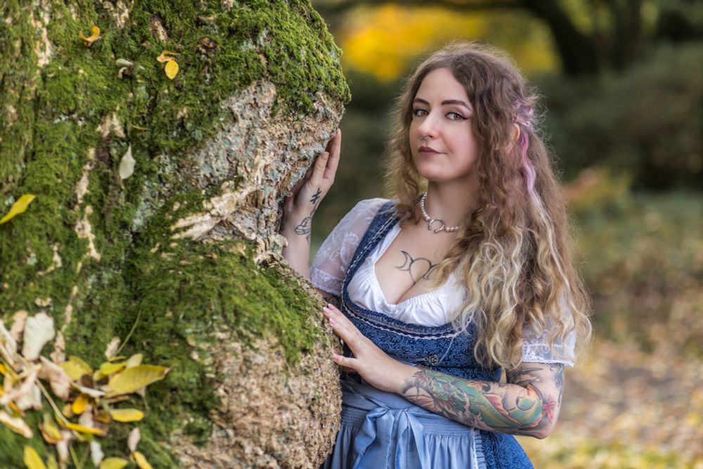 Eine Person mit langen, welligen braunen Haaren, die violette Strähnen haben, steht in einem herbstlichen Wald. Sie trägt ein traditionelles blaues Dirndl mit weißer Bluse, die mit Spitze verziert ist, und hat auffällige Tattoos auf den Armen und dem Dekolleté, darunter ein Symbol mit drei Monden und andere Designs. Die Person lehnt mit einer Hand an einem moosbewachsenen Baumstamm, während bunte Herbstblätter am Boden und im Hintergrund zu sehen sind. Der Hintergrund zeigt ein malerisches Laub in Gelb- und Orangetönen sowie grüne Bäume.