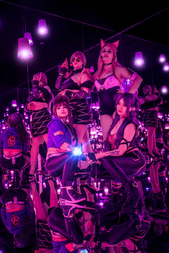 Vier Frauen posieren in auffälligen, futuristischen Outfits in einem Raum voller Spiegel. Die Szene ist in neonlila Licht getaucht, das von den zahlreichen Hängelampen reflektiert wird. Jede der Frauen trägt ein einzigartiges Cosplay, inspiriert von einem Musik- oder Gaming-Universum. Eine Frau mit Katzenohren steht mit selbstbewusstem Blick, während eine andere mit einer leuchtenden Kugel in der Hand kniet. Zwei weitere Figuren stehen mit coolen Posen im Hintergrund. Der Raum erzeugt durch die Spiegelung eine endlose Illusion, als würden sie in einer glitzernden Cyberwelt schweben.