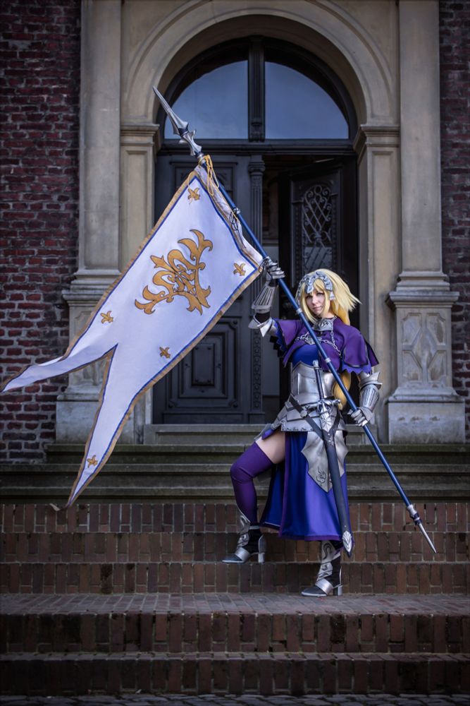 Eine Person in einer eindrucksvollen Ritterrüstung posiert auf einer Steintreppe vor einem historischen Gebäude. Sie trägt silberne Rüstungsteile über einem langen, königsblauen Gewand mit violetten Akzenten. Eine blonde Perücke mit einem Stirnband rundet das majestätische Erscheinungsbild ab. In der rechten Hand hält sie eine große, weiße Fahne mit goldenen Lilien und Verzierungen, die im Wind weht. Mit entschlossenem Blick und einer stolzen Haltung verkörpert sie eine heldenhafte Kriegerfigur, die an eine mittelalterliche Jeanne d’Arc erinnert.